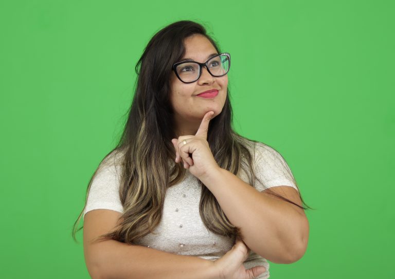 Mulher de cabelos longos, usando óculos e camiseta branca, em frente a um fundo verde. Postura pensativa, olhando para cima e encostando o dedo indicador no queixo.
